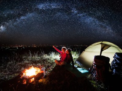Night camping near the town. Young couple sitting near campfire and tent, looking at beautiful night sky full of stars and enjoying night scene. Woman is pointing at the sky. Astrophotography
